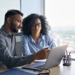 Indian male ceo executive manager mentor giving consultation on financial operations to female African American colleague intern using laptop sitting in modern office near panoramic window.